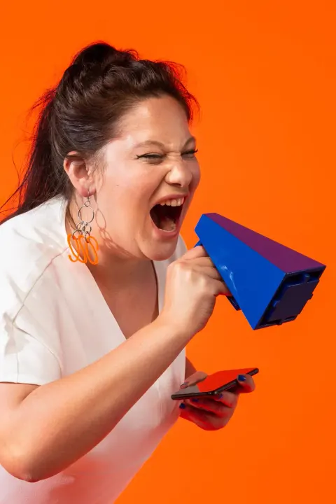 A woman screams into Akrasia’s blue megaphone, phone in hand, against an orange backdrop
