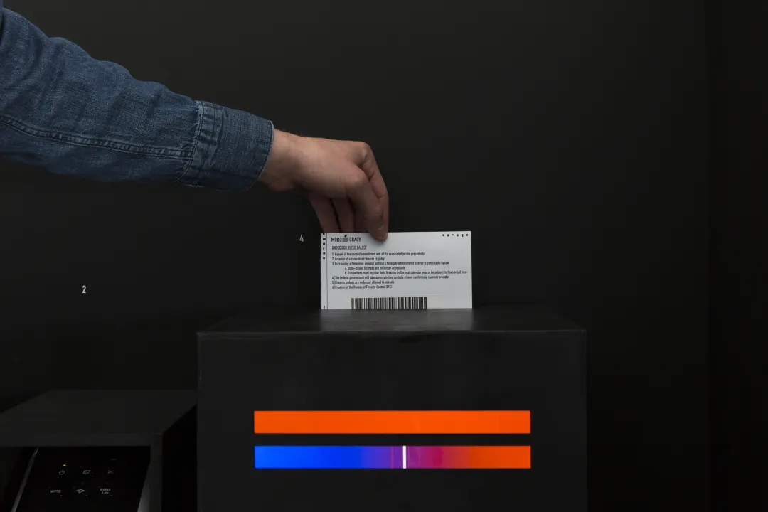A ballot with a printed sticker going into the box, orange and blue lights below